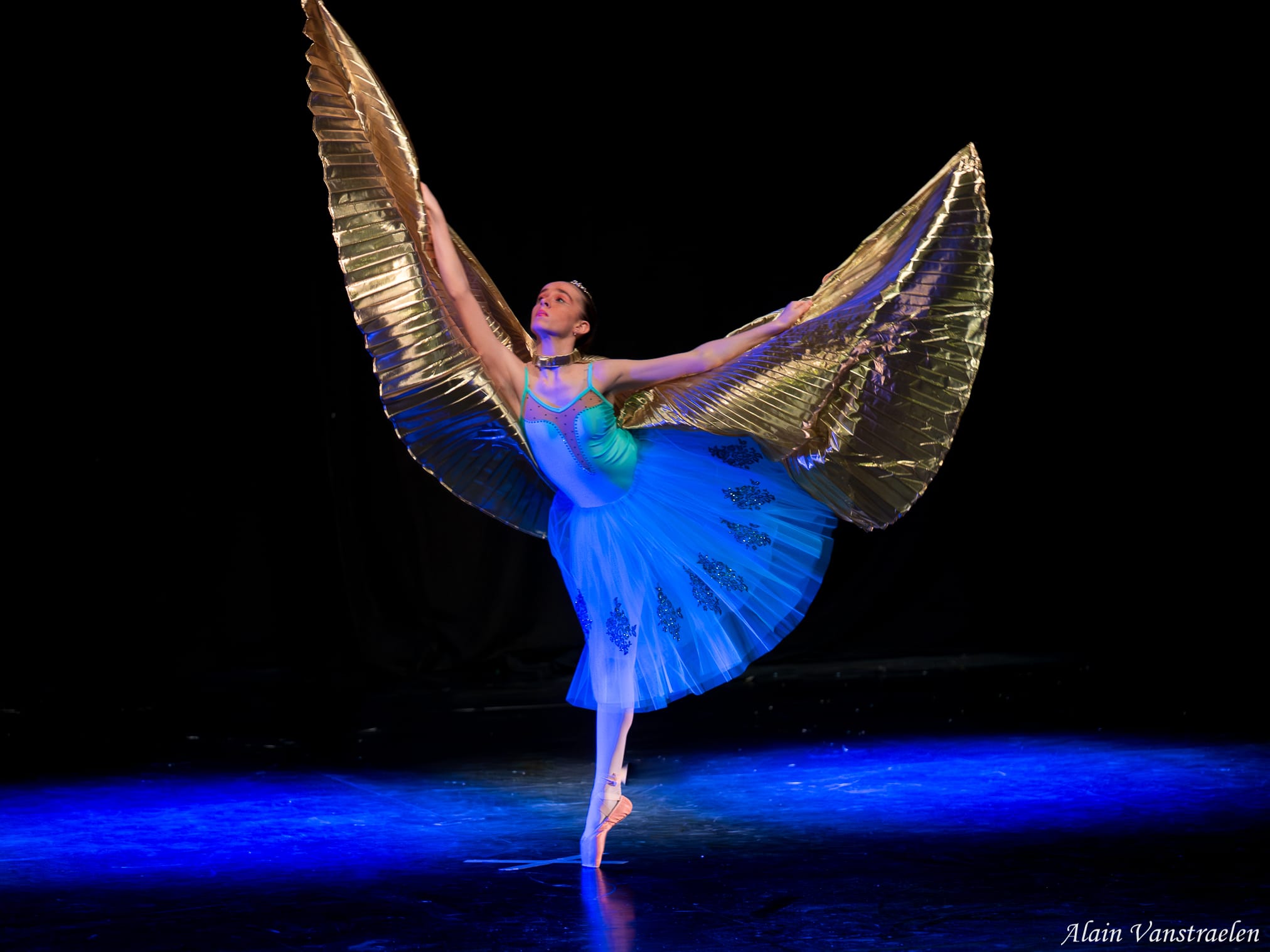 Danseuse classique sur pointes en tutu bleu, de grandes ailes dorées déployées, sur la scène du Concours
