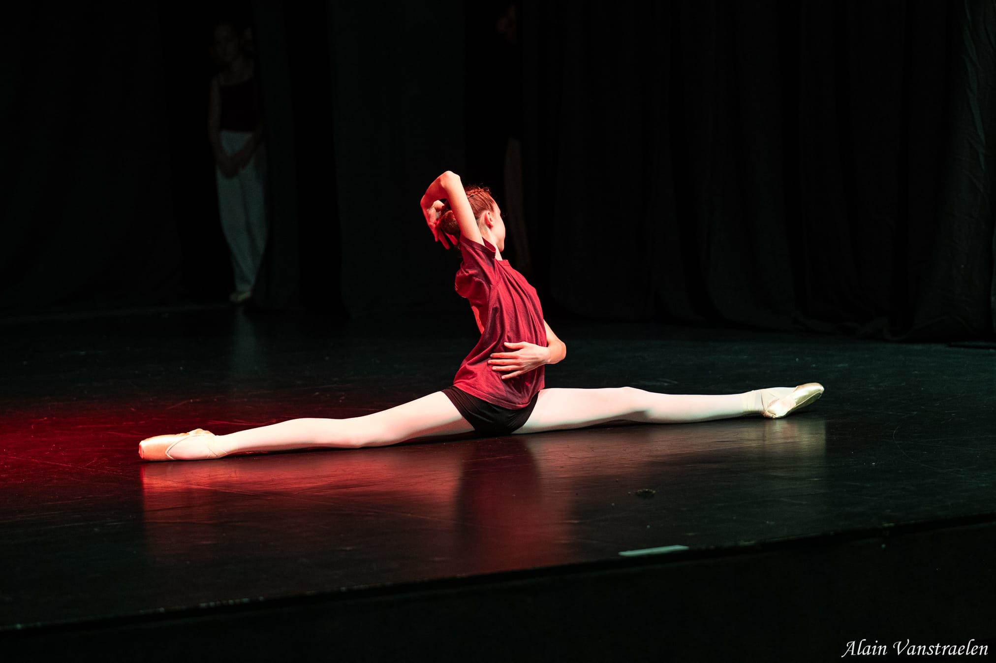 Danseuse sur pointes en grand écart facial au sol, lumière rouge rasante